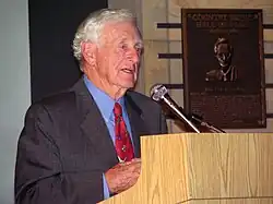 White-haired elderly gentleman in suit and tie speaks at a podium.